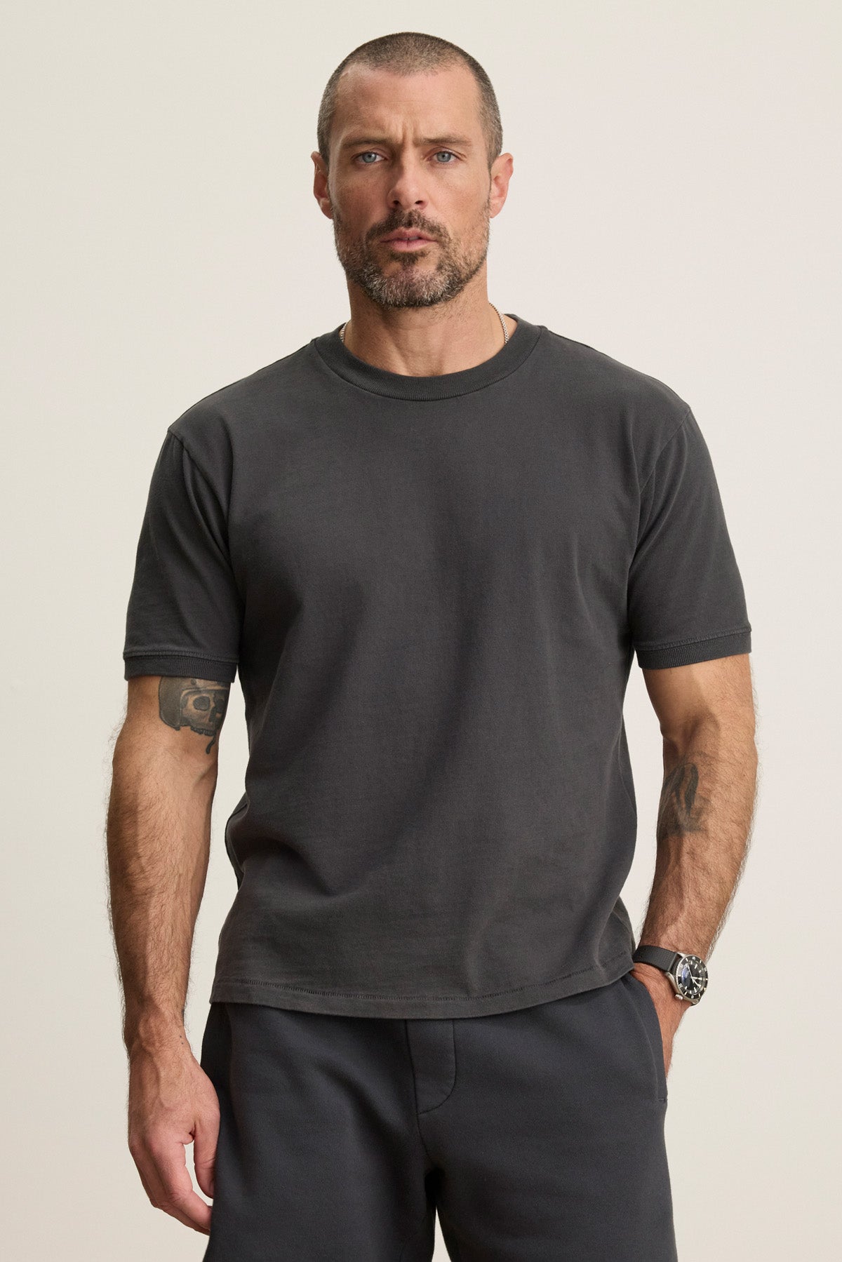A man with short hair and a beard wears the ELIAS TEE by Velvet by Graham & Spencer—a dark grey, heavy jersey crew neck t-shirt—with pants. He stands against a plain light background, showing tattoos on his left arm and a watch on his right wrist.-40606035869889