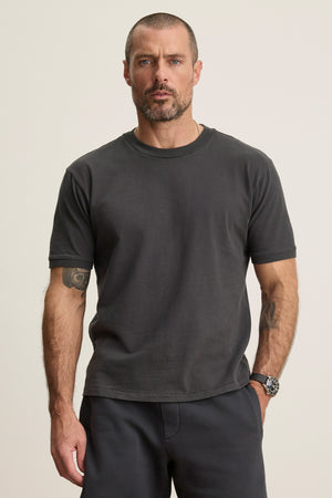 A man with short hair and a beard wears the ELIAS TEE by Velvet by Graham & Spencer—a dark grey, heavy jersey crew neck t-shirt—with pants. He stands against a plain light background, showing tattoos on his left arm and a watch on his right wrist.
