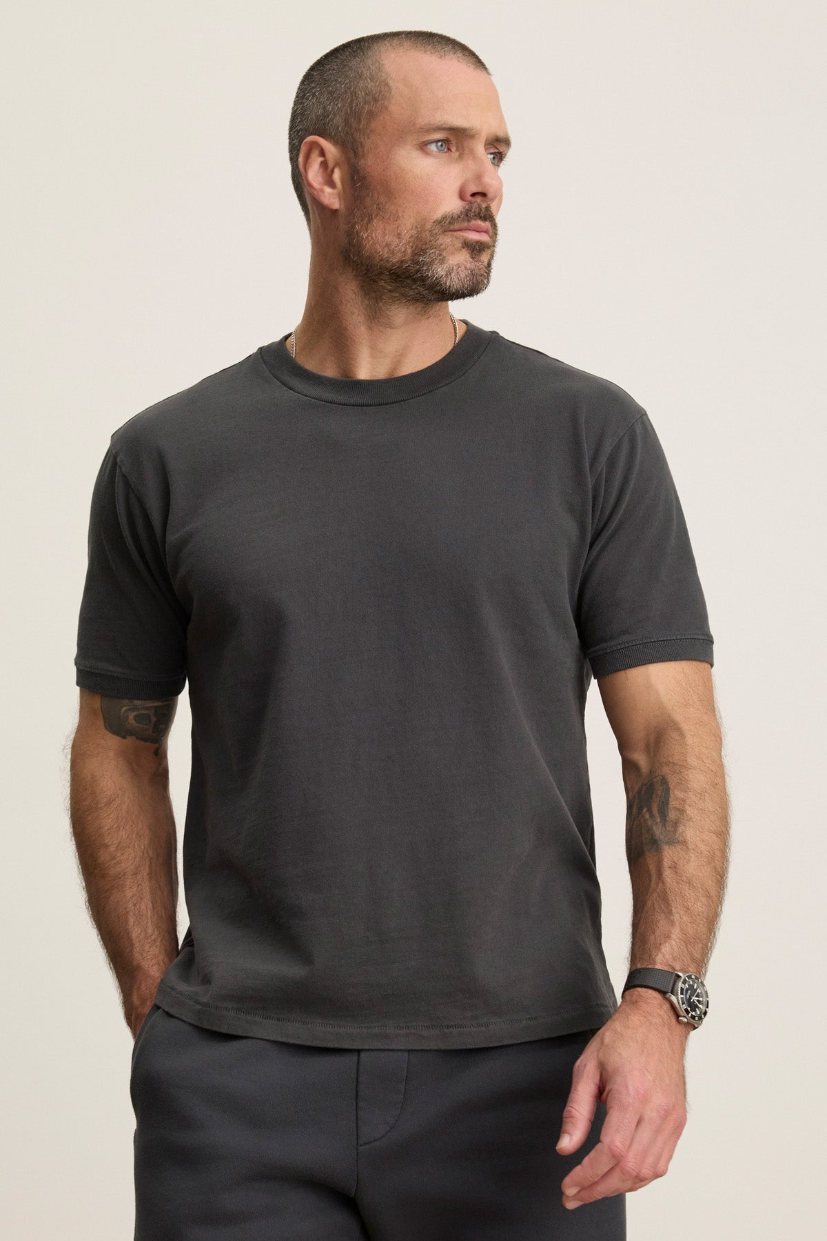 A man with short hair and a beard stands with his hands in his pockets, wearing the ELIAS TEE by Velvet by Graham & Spencer—a plain black heavy jersey crewneck t-shirt—paired with dark pants, looking to the side against a plain background.-40606035837121