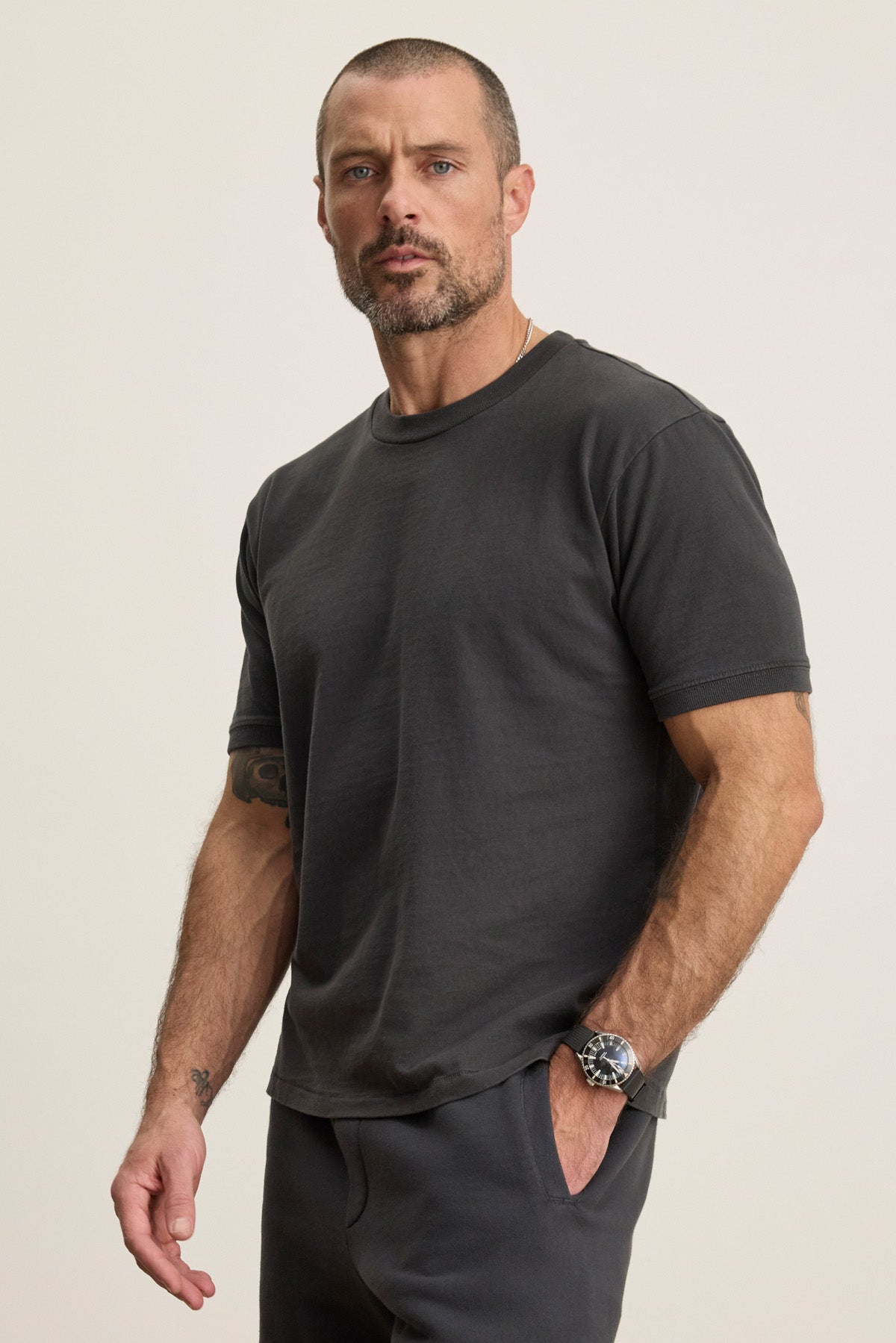 A man with short hair and a beard wears the ELIAS TEE by Velvet by Graham & Spencer—a plain dark heavy jersey crewneck t-shirt—paired with dark pants, standing with one hand in his pocket against a neutral background.