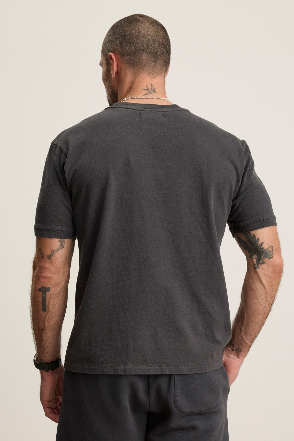 A man with short hair, tattoos, and a necklace is seen from behind wearing the ELIAS TEE by Velvet by Graham & Spencer—a plain dark gray heavy jersey crewneck t-shirt paired with matching pants for an everyday staple look.