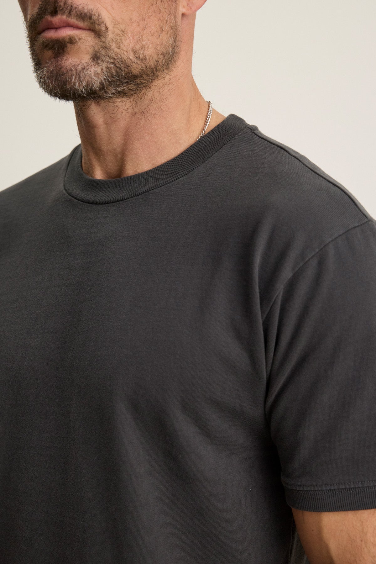 A man with a trimmed beard wears the ELIAS TEE by Velvet by Graham & Spencer, a plain dark gray heavy jersey crewneck t-shirt, paired with a thin chain necklace—an everyday staple look shown from the shoulders up.