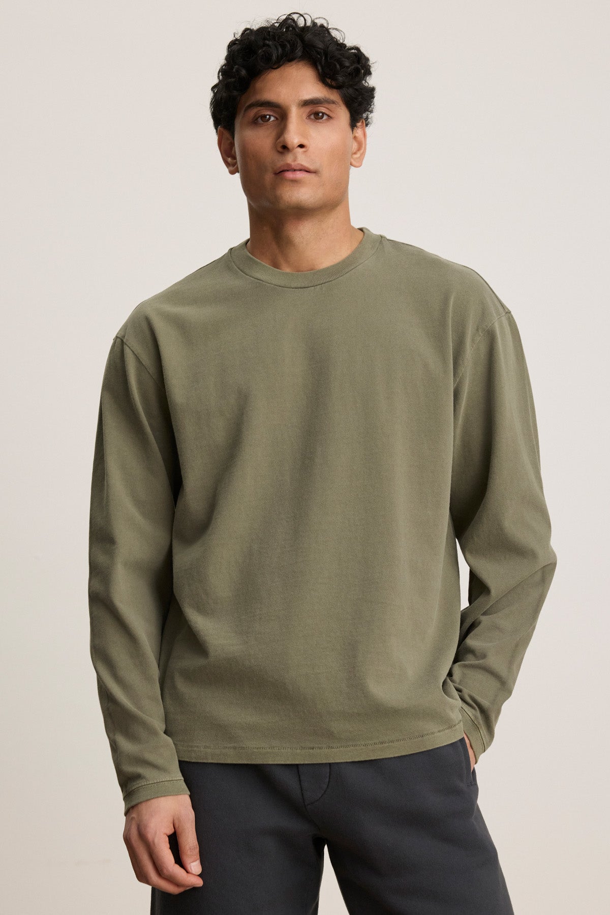 A man with short curly hair wears the FINNICK TEE by Velvet by Graham & Spencer, an olive green heavyweight jersey with a ribbed crew neckline, paired with dark pants against a plain light background.-40452482597057