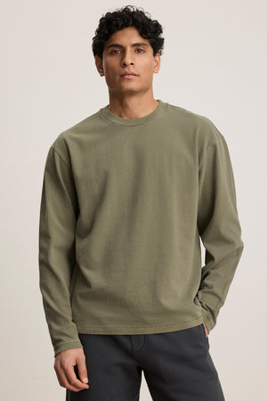 A man with short curly hair wears the FINNICK TEE by Velvet by Graham & Spencer, an olive green heavyweight jersey with a ribbed crew neckline, paired with dark pants against a plain light background.