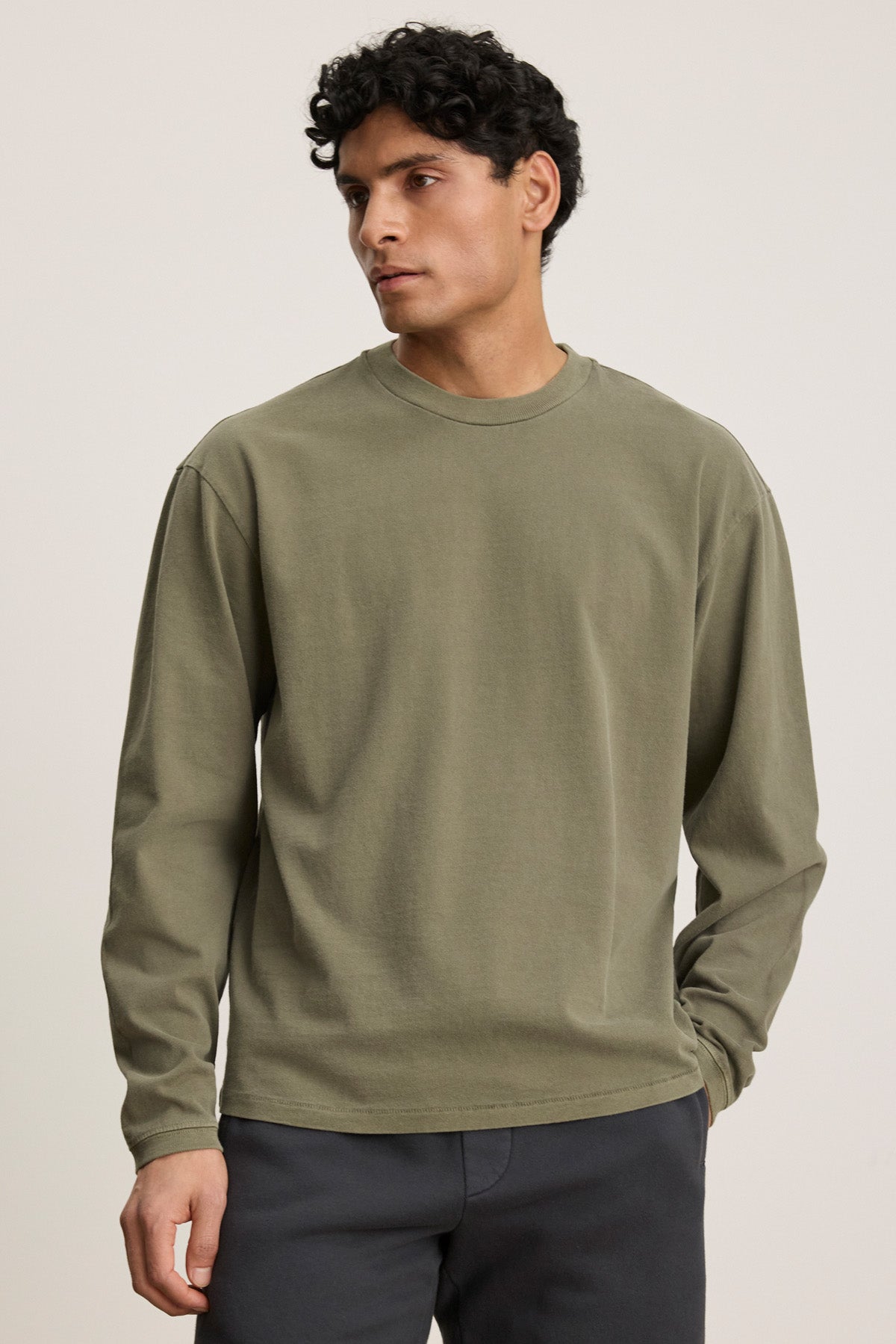 A man wears the FINNICK TEE by Velvet by Graham & Spencer, an olive green heavyweight jersey with a ribbed crew neckline, paired with dark pants against a plain light background.-40452482564289