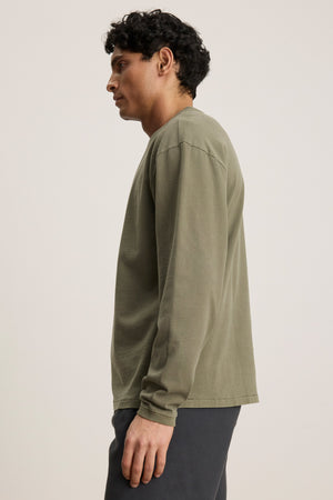 A man in profile wears the Velvet by Graham & Spencer FINNICK TEE in olive green—a heavyweight jersey with a ribbed crew neckline—paired with dark pants, set against a plain light background.