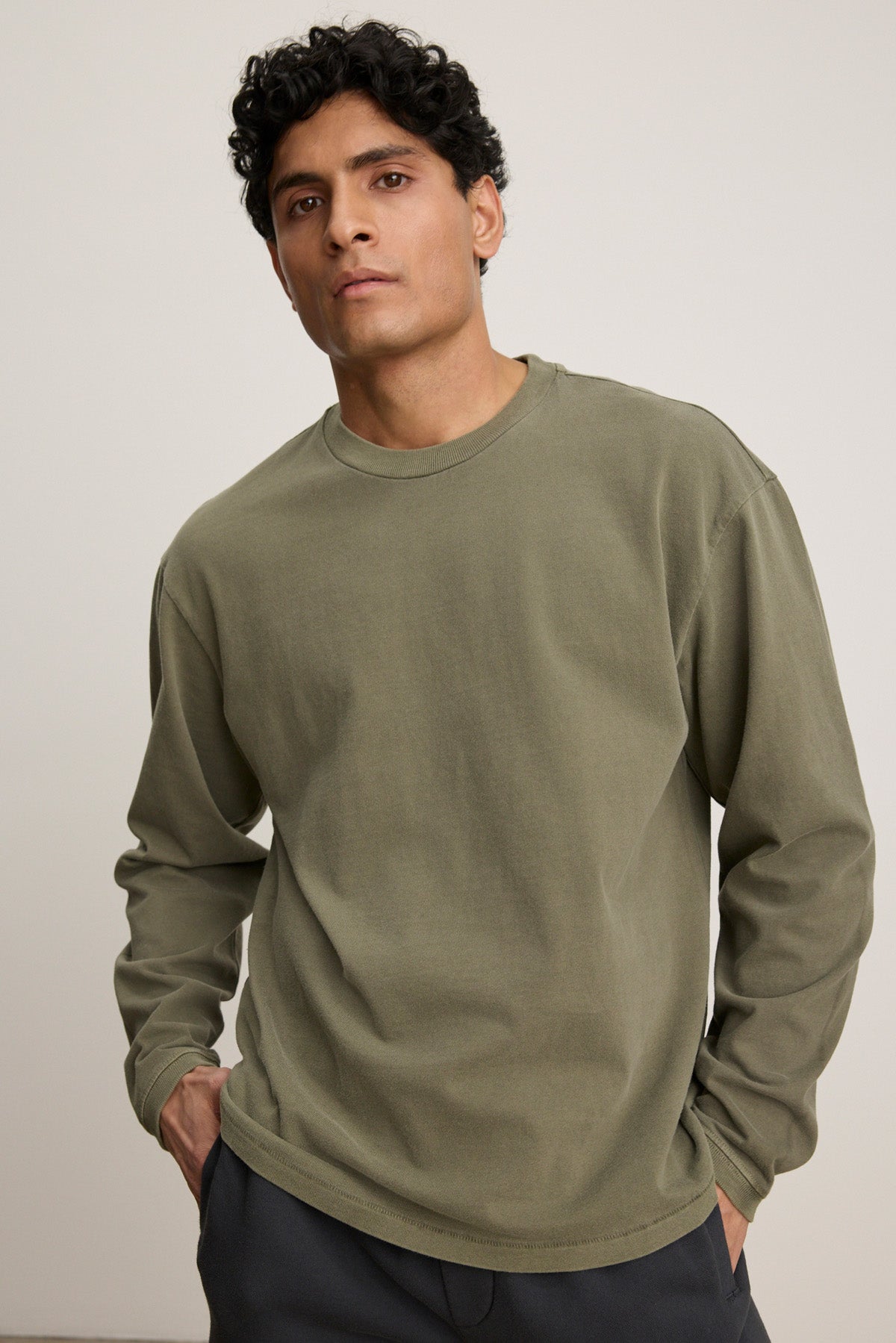 A person models the FINNICK TEE by Velvet by Graham & Spencer, an olive green heavyweight jersey tee with a ribbed crew neckline, standing against a plain light background with hands in pockets.-40452482400449