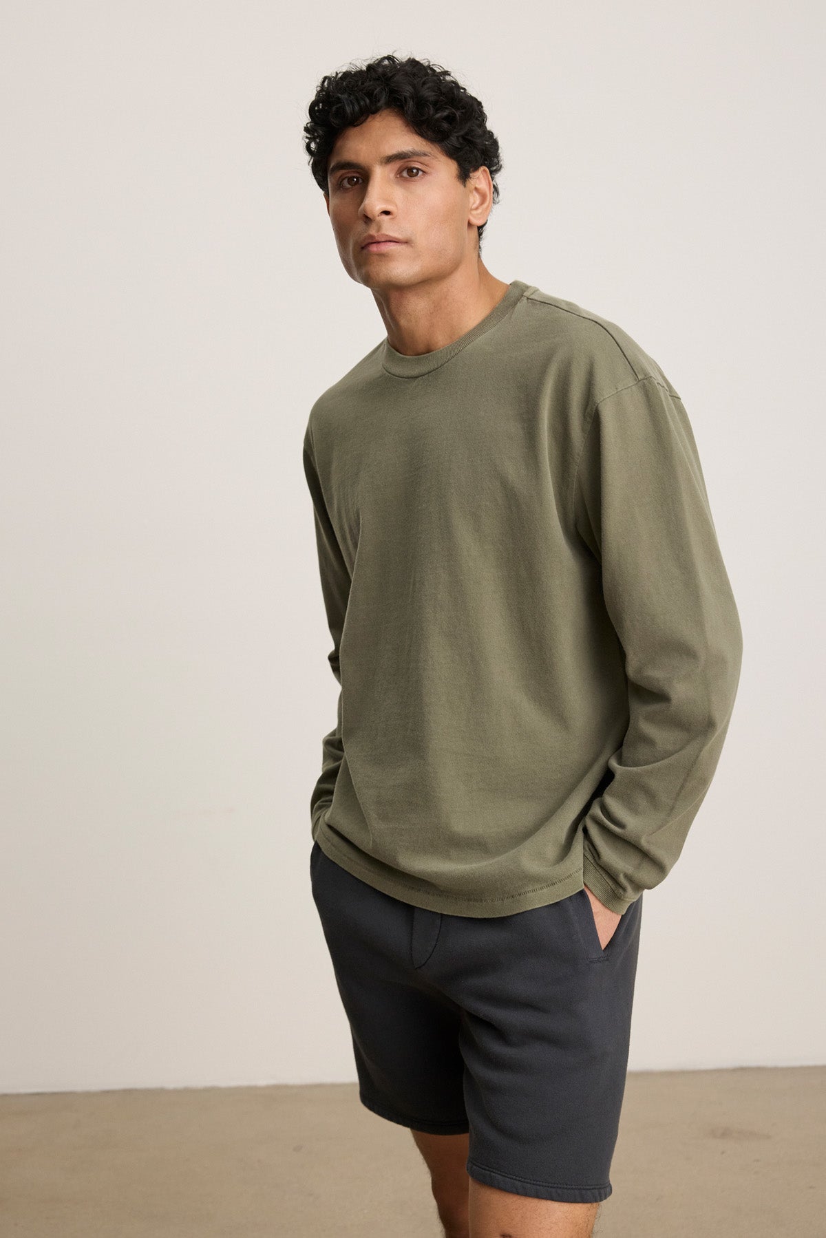 A man stands indoors with his hands in his pockets, wearing the FINNICK TEE by Velvet by Graham & Spencer—an olive green heavyweight jersey crewneck tee—and dark shorts, looking at the camera.
