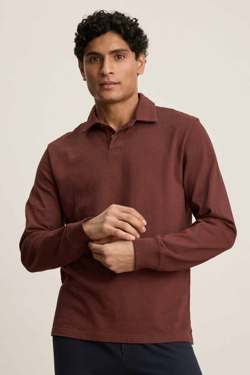 A man with short curly hair wears the HAWKINS POLO by Velvet by Graham & Spencer in maroon midweight jersey and dark pants, standing against a plain light background.