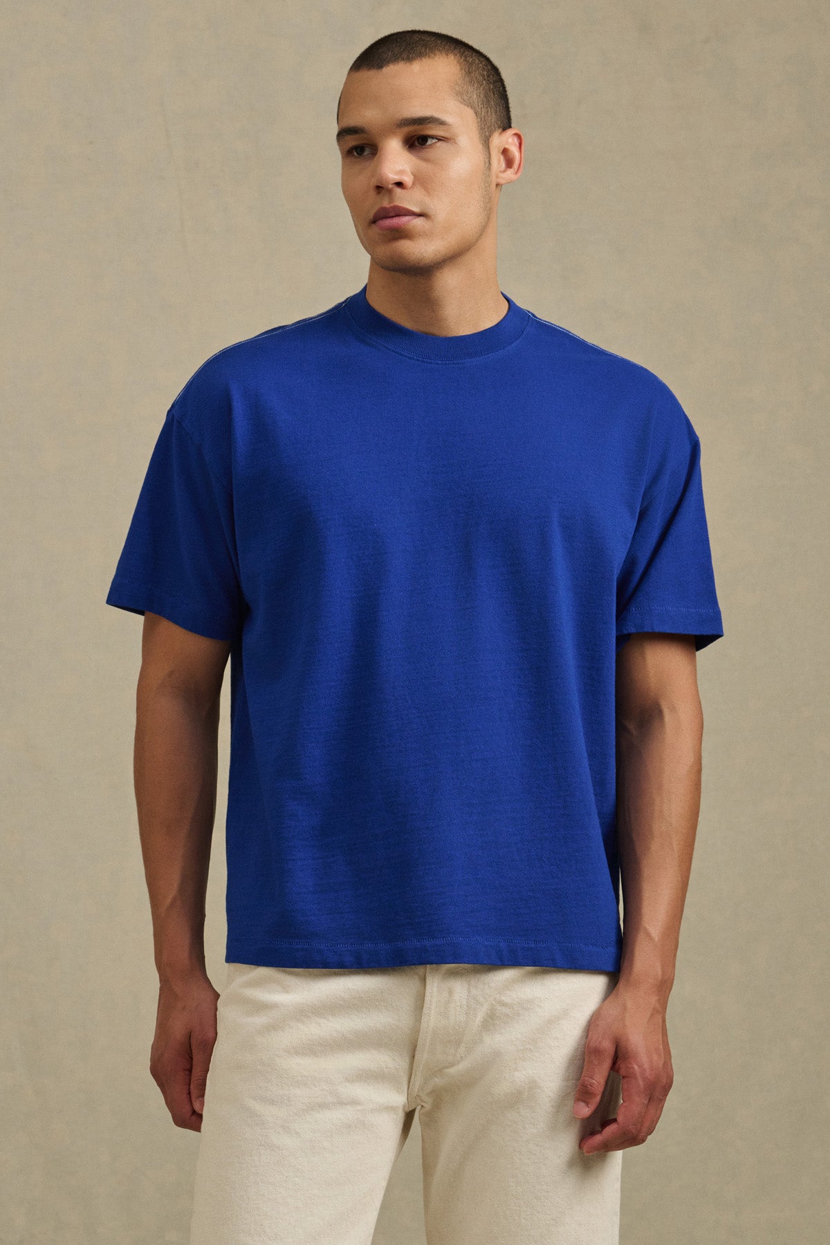 A person stands against a plain background wearing the ISAAC TEE by Velvet by Graham & Spencer in royal blue, featuring a boxy relaxed fit and paired with off-white pants.-41338468827329