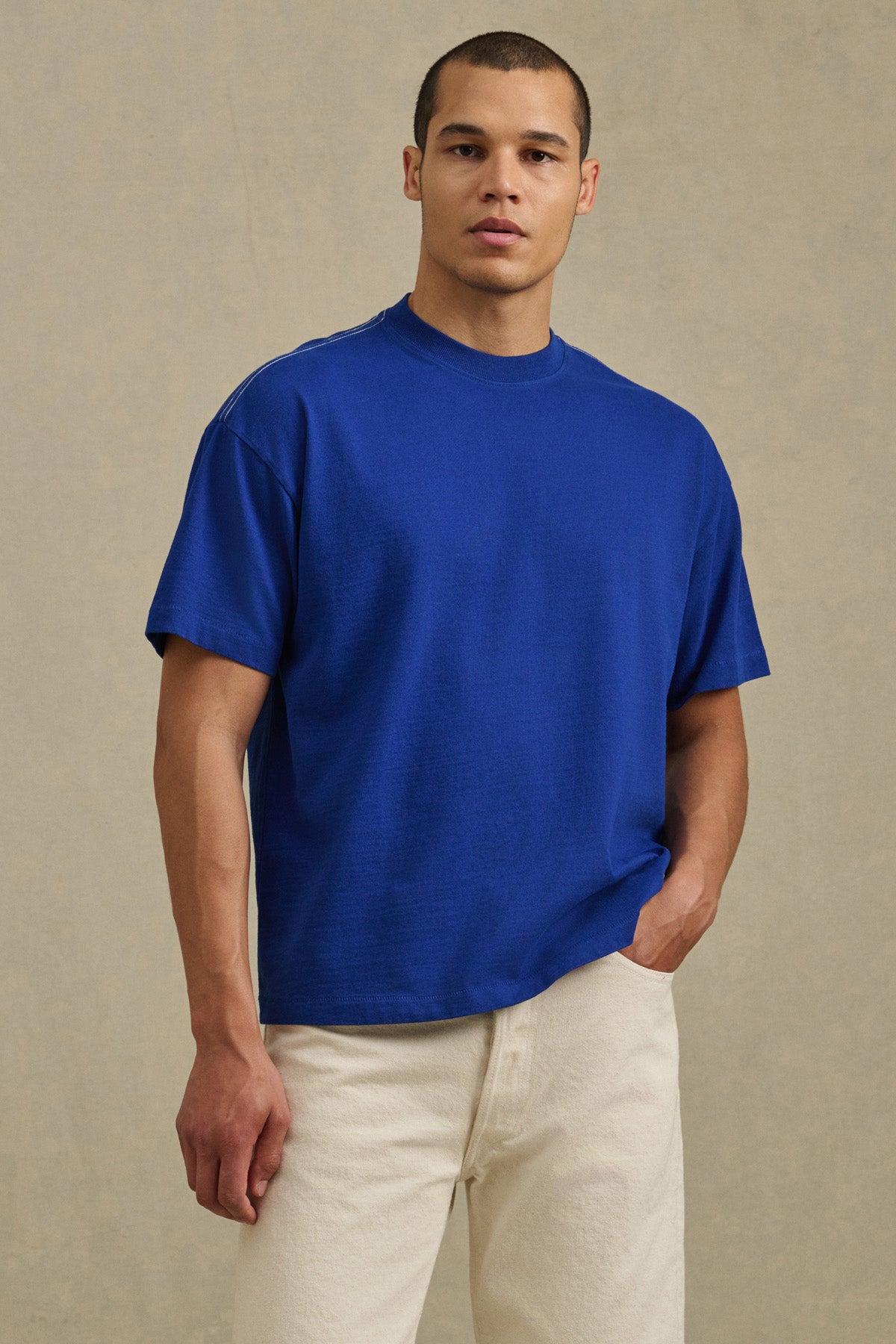 A person stands against a neutral background wearing the Velvet by Graham & Spencer ISAAC TEE in blue heavyweight cotton jersey with off-white pants, looking at the camera with one hand in their pocket.