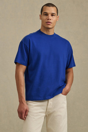 A person stands against a neutral background wearing the Velvet by Graham & Spencer ISAAC TEE in blue heavyweight cotton jersey with off-white pants, looking at the camera with one hand in their pocket.