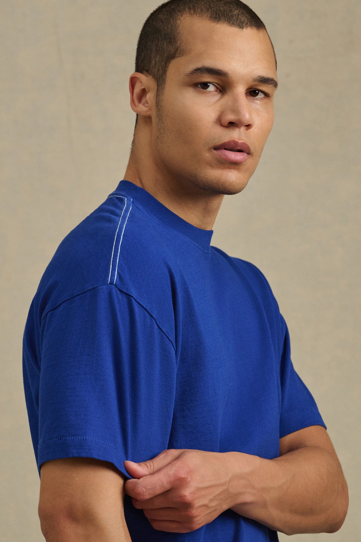 A person with short hair wears the ISAAC TEE by Velvet by Graham & Spencer—a blue, heavier-weight cotton jersey with contrast stitching—while looking toward the camera against a plain background.-41338468761793