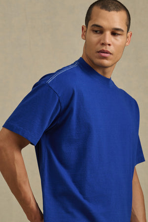 A man with short hair wears the Velvet by Graham & Spencer ISAAC TEE, a relaxed fit blue tee crafted from heavier-weight cotton jersey, as he stands against a neutral background looking to his left.
