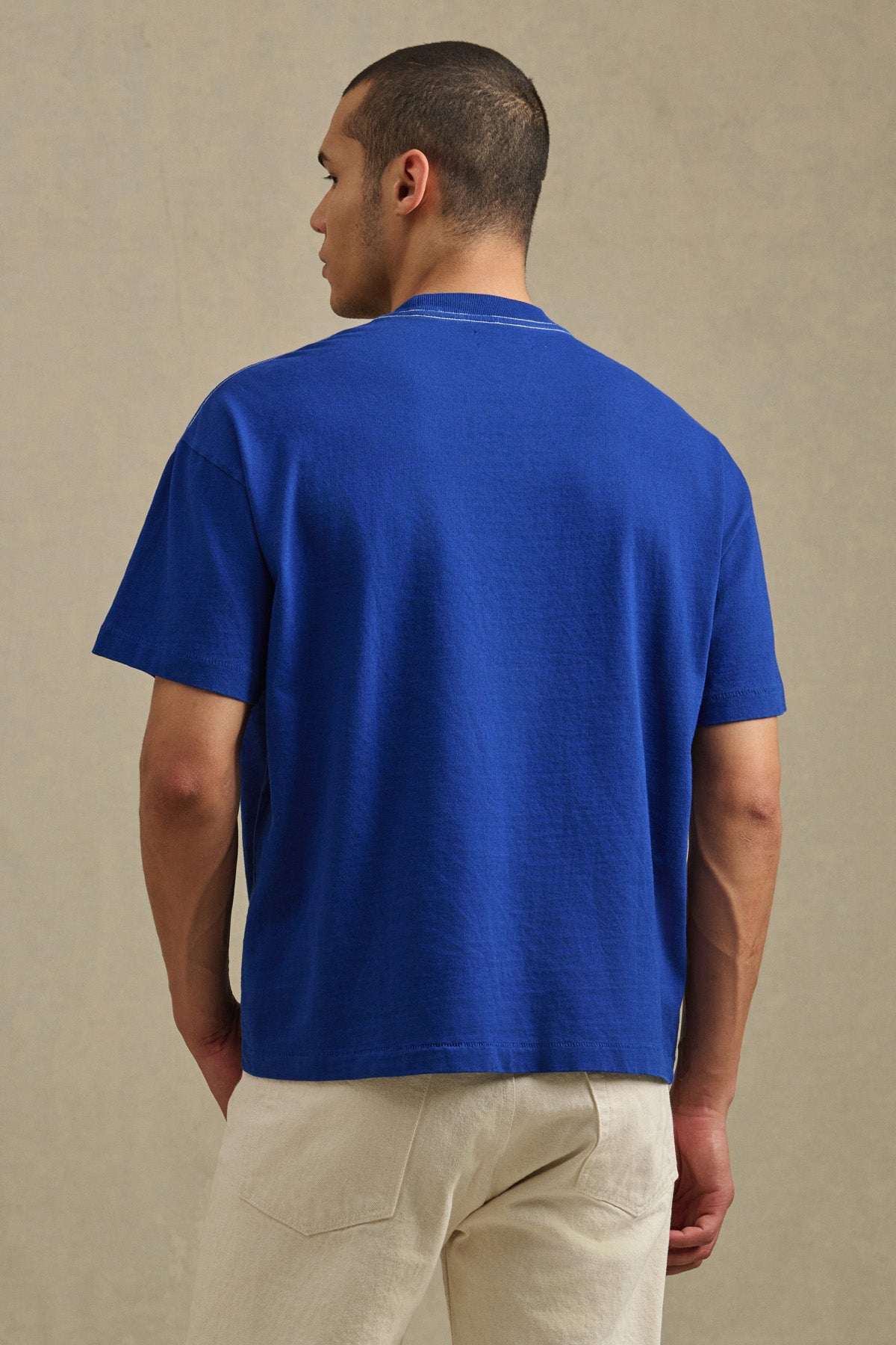 A person stands facing away, wearing the ISAAC TEE by Velvet by Graham & Spencer in plain blue and light-colored pants against a neutral background.-41338468663489