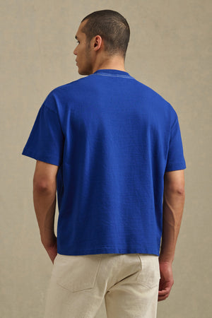A person stands facing away, wearing the ISAAC TEE by Velvet by Graham & Spencer in plain blue and light-colored pants against a neutral background.