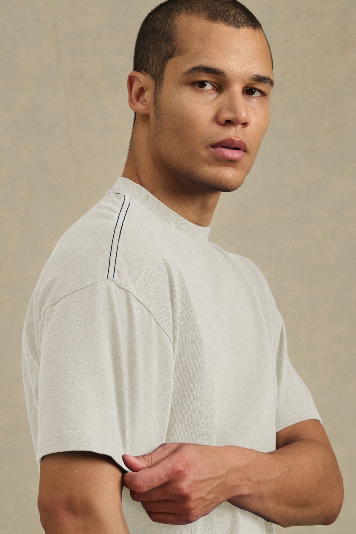 A person with short hair wears the Velvet by Graham & Spencer ISAAC TEE, a boxy relaxed-fit light short-sleeve shirt with a dark shoulder stripe, standing against a plain beige background.-41105022189761