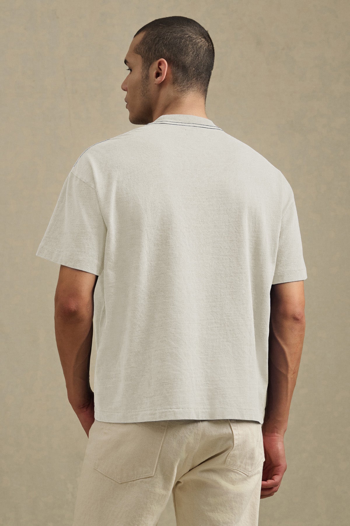 A man faces away wearing the ISAAC TEE by Velvet by Graham & Spencer in light gray, featuring a boxy relaxed fit with short sleeves, paired with beige pants against a neutral background.-41105022156993