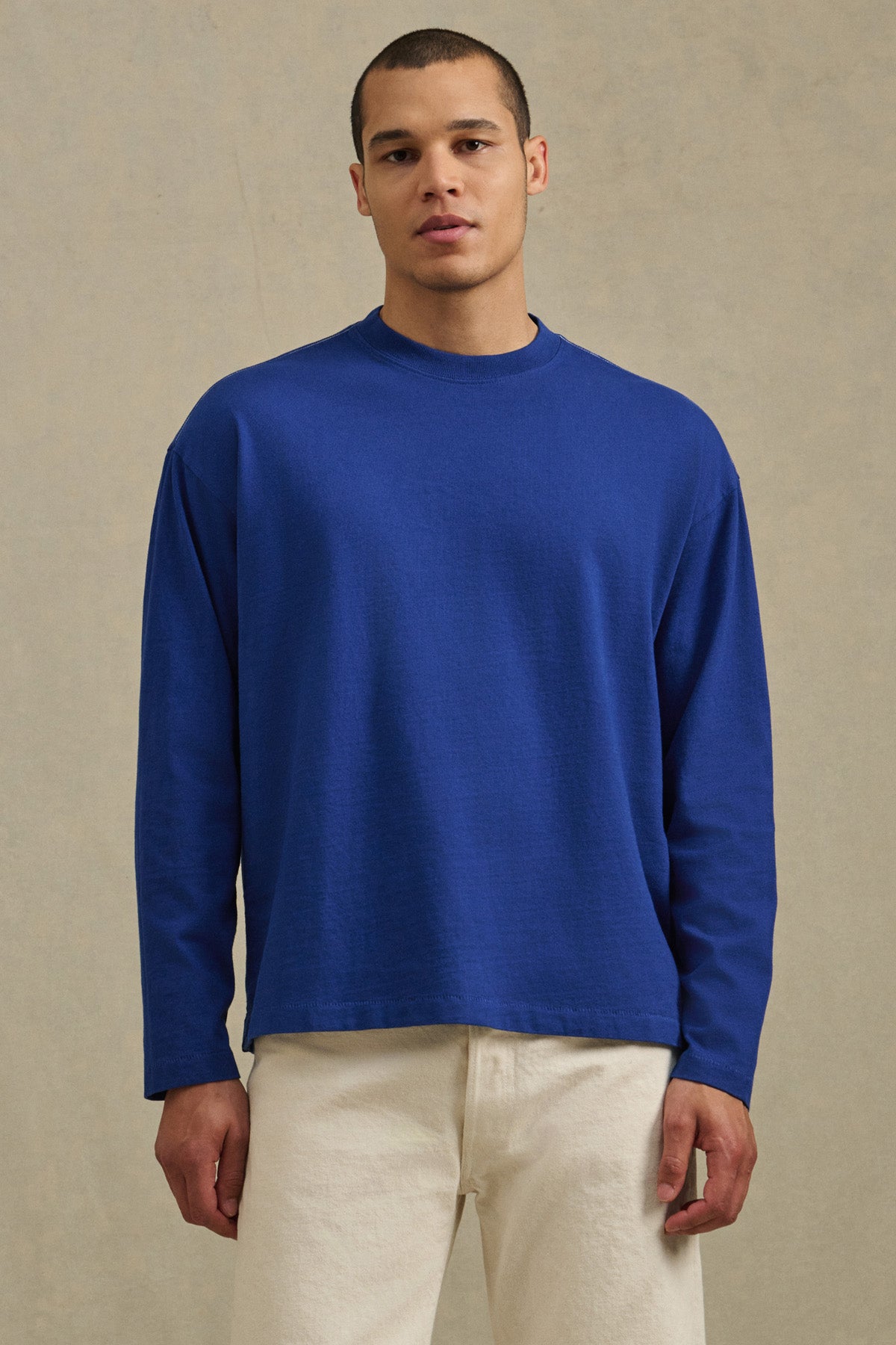 A man wearing the Velvet by Graham & Spencer THEO TEE in blue and off-white pants stands against a plain beige background.-41338474168513
