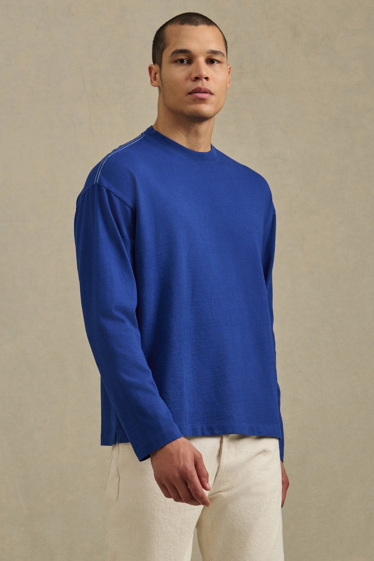 A person stands against a neutral background wearing the relaxed fit, long-sleeved blue THEO TEE by Velvet by Graham & Spencer with off-white pants, looking directly at the camera.