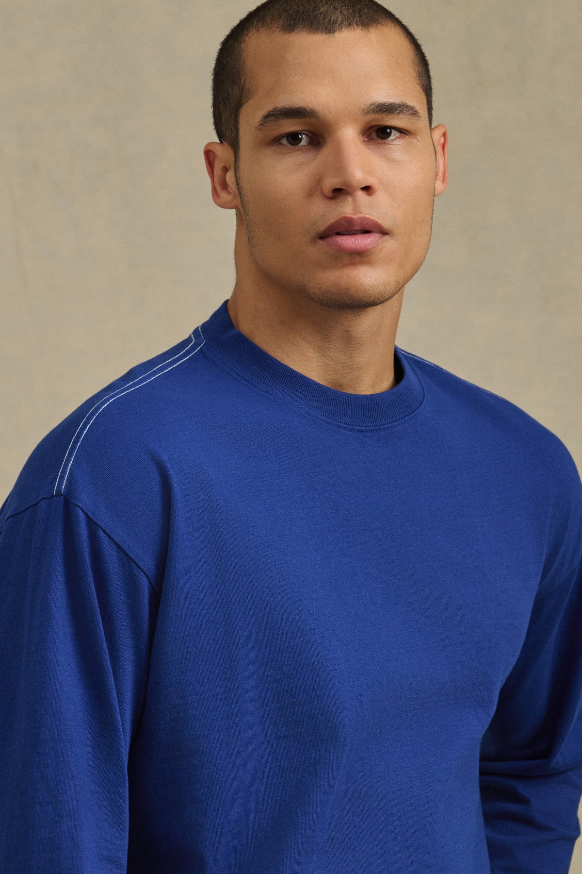 Someone with short hair wears the THEO TEE by Velvet by Graham & Spencer, a blue heavyweight long-sleeve jersey shirt, standing against a neutral background and gazing slightly to the side.