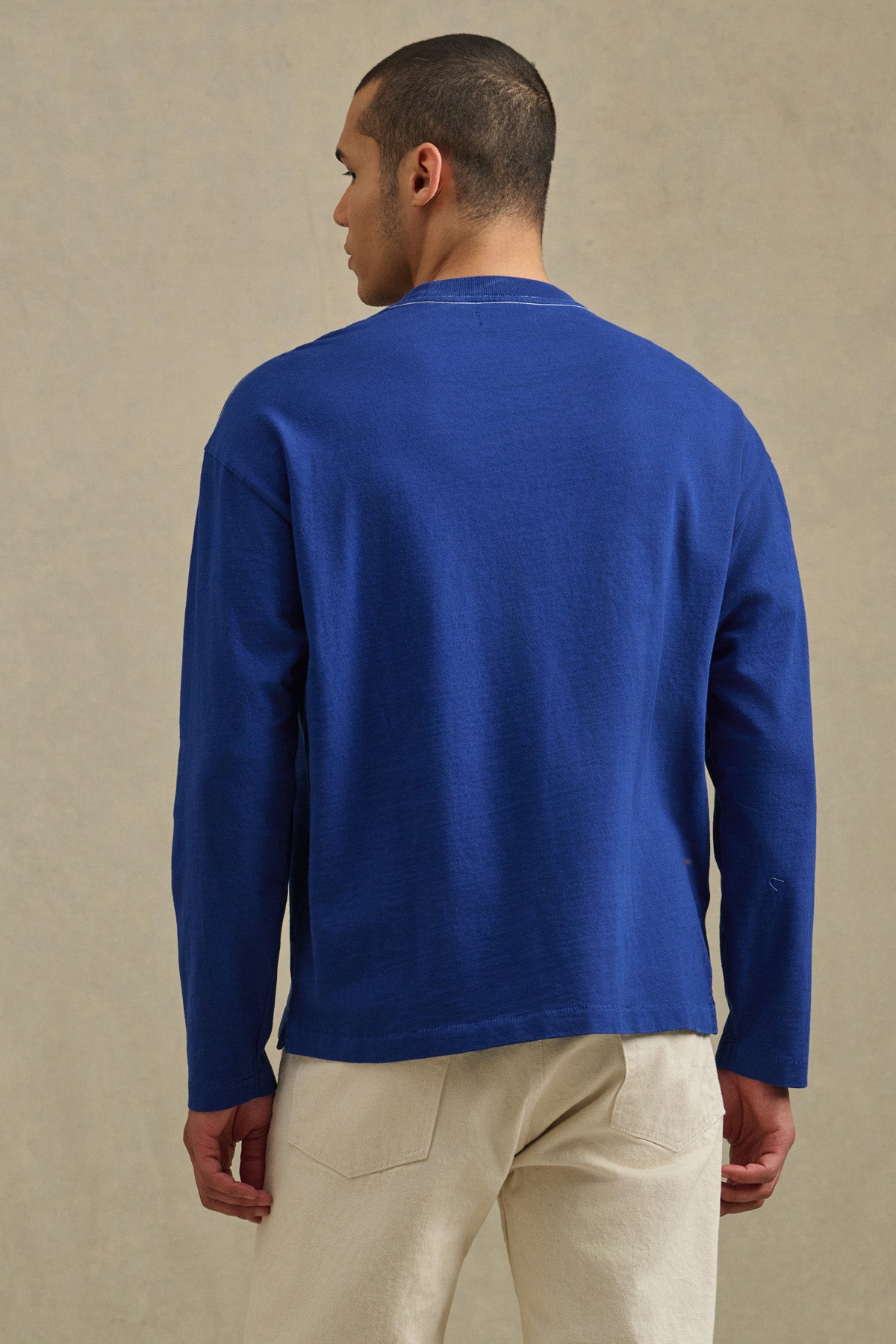A man with short hair stands with his back to the camera, wearing the Velvet by Graham & Spencer THEO TEE—a relaxed fit, long-sleeve blue shirt—paired with light pants against a neutral background.