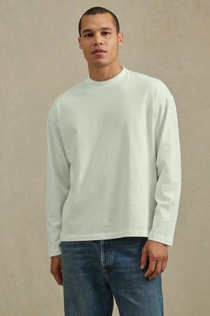 A person faces the camera, wearing the Velvet by Graham & Spencer THEO TEE—a relaxed fit, heavyweight jersey long-sleeve shirt—with blue jeans against a neutral background.