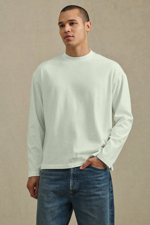 A man wears the Velvet by Graham & Spencer THEO TEE, a relaxed-fit heavier-weight jersey classic tee, with blue jeans against a neutral background.