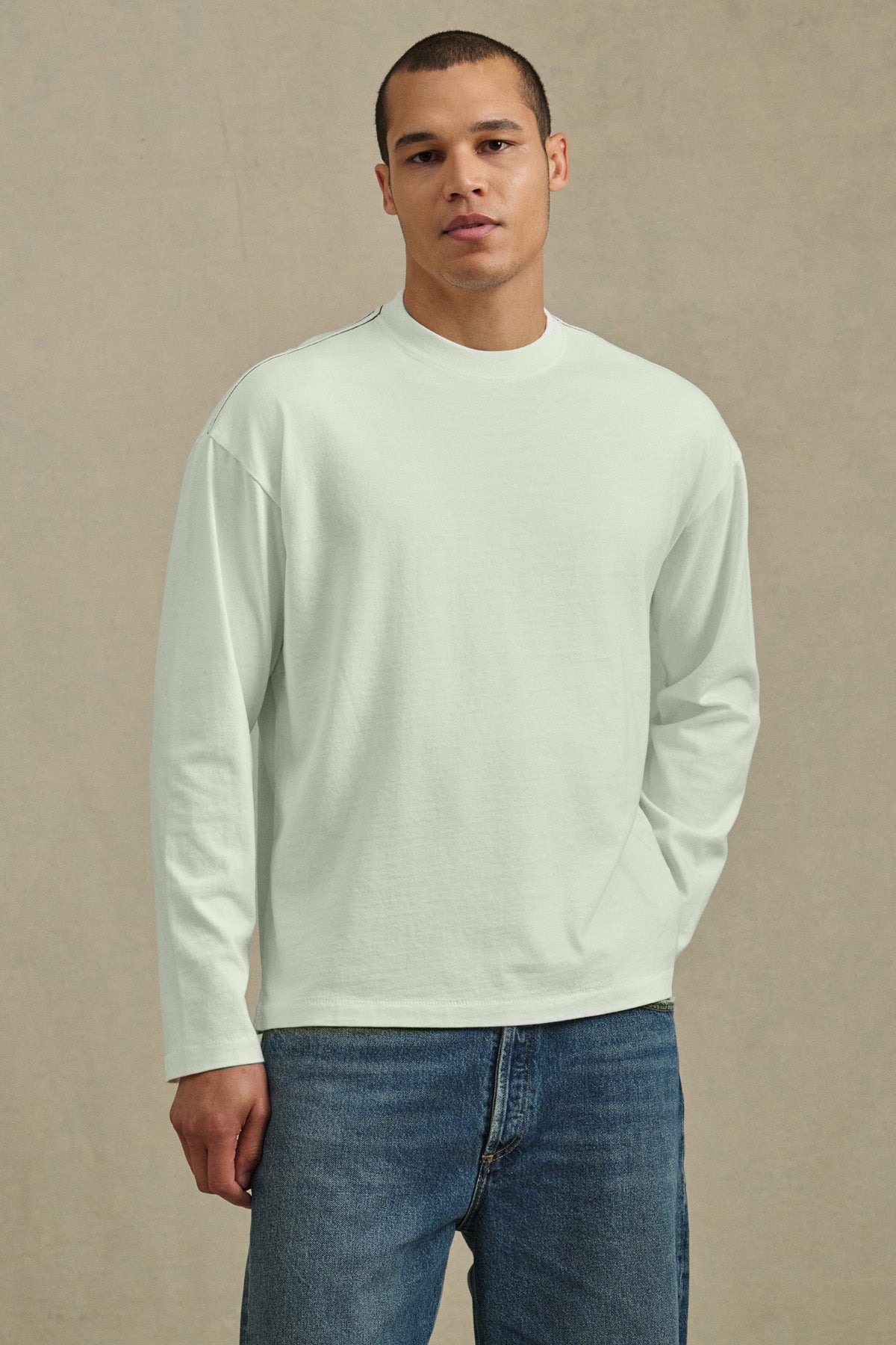 A person stands against a neutral background wearing Velvet by Graham & Spencer's THEO TEE, a relaxed fit, heavier-weight jersey long-sleeve white shirt, paired with blue jeans, looking directly at the camera.-41105046569153