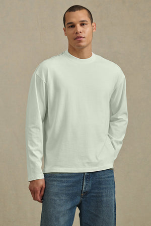 A person stands against a neutral background wearing Velvet by Graham & Spencer's THEO TEE, a relaxed fit, heavier-weight jersey long-sleeve white shirt, paired with blue jeans, looking directly at the camera.