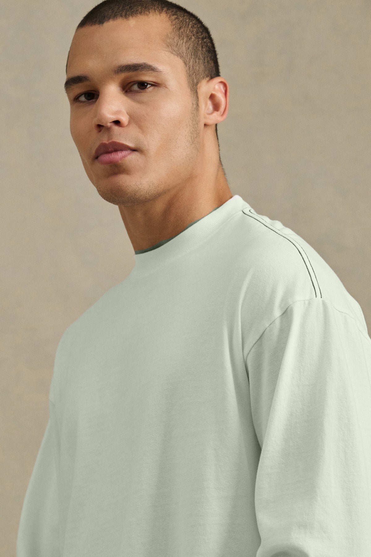 A man with short hair wears the relaxed fit THEO TEE by Velvet by Graham & Spencer, a light-colored long-sleeve shirt, and looks toward the camera against a plain, neutral background.