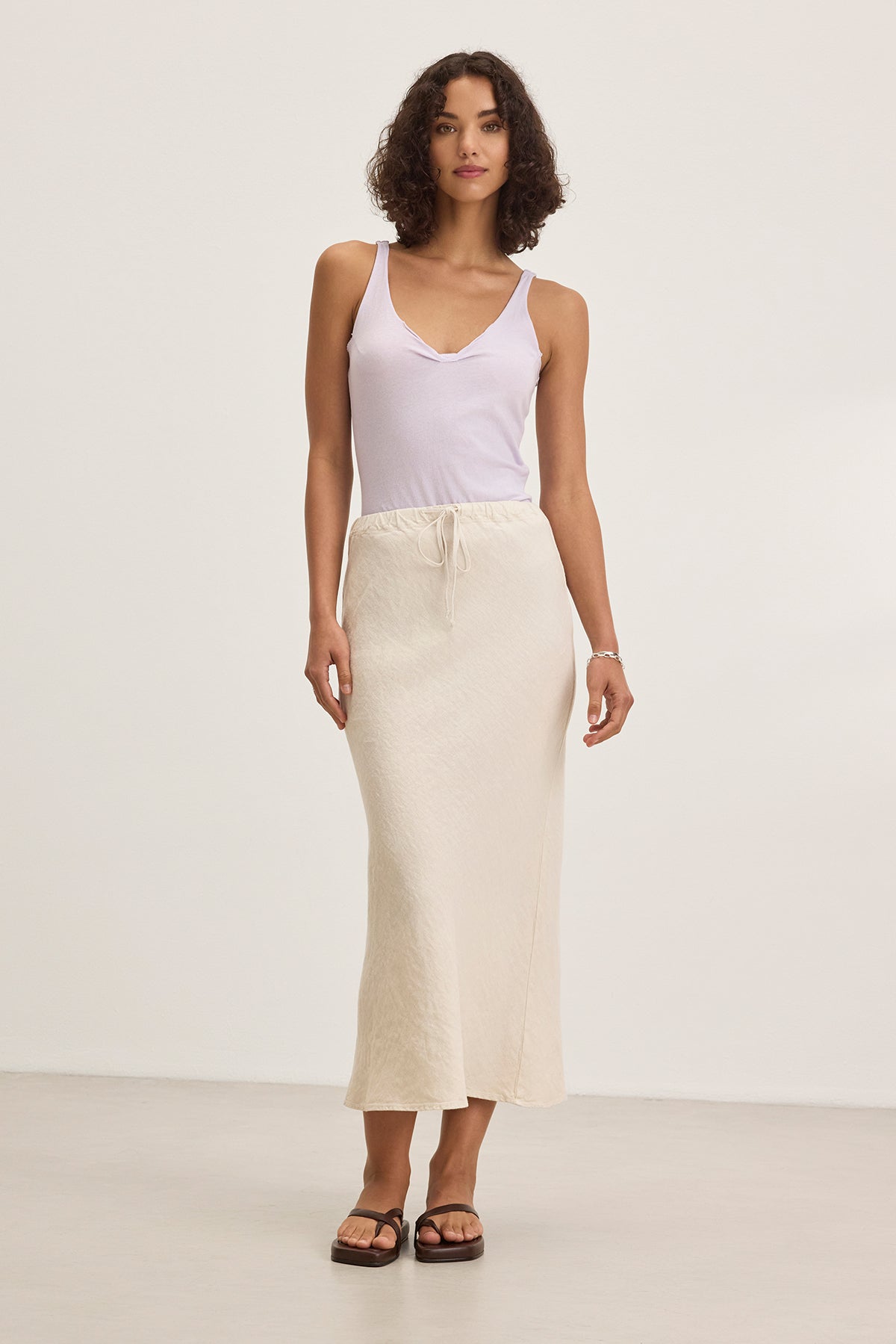 A person with curly hair models a white tank top, the BEKA HEAVY LINEN SKIRT by Velvet by Graham & Spencer in cream, and black sandals against a plain white background.-38662641254593