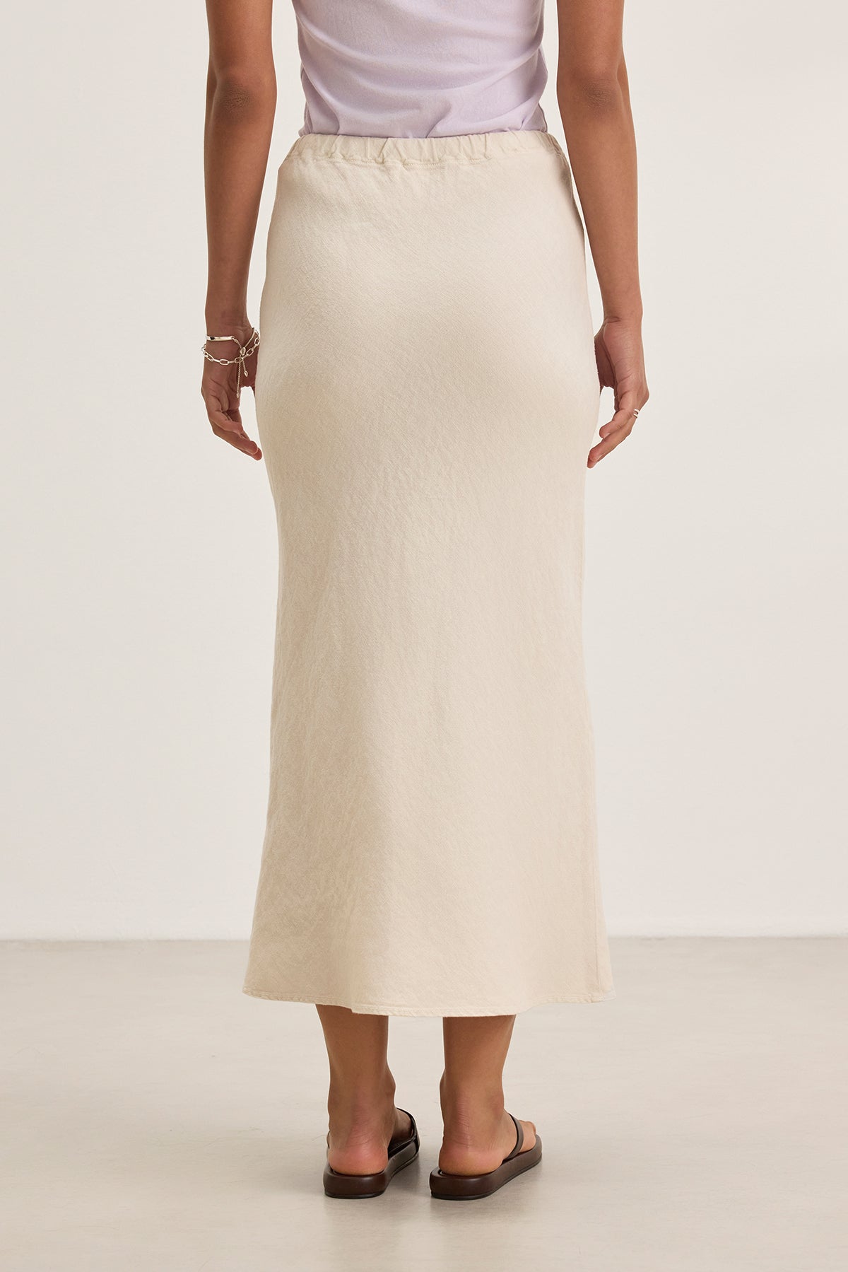 Back view of a person wearing a white top and the BEKA HEAVY LINEN SKIRT by Velvet by Graham & Spencer in beige, bias cut maxi style, paired with black sandals, standing against a plain background.-38662641189057