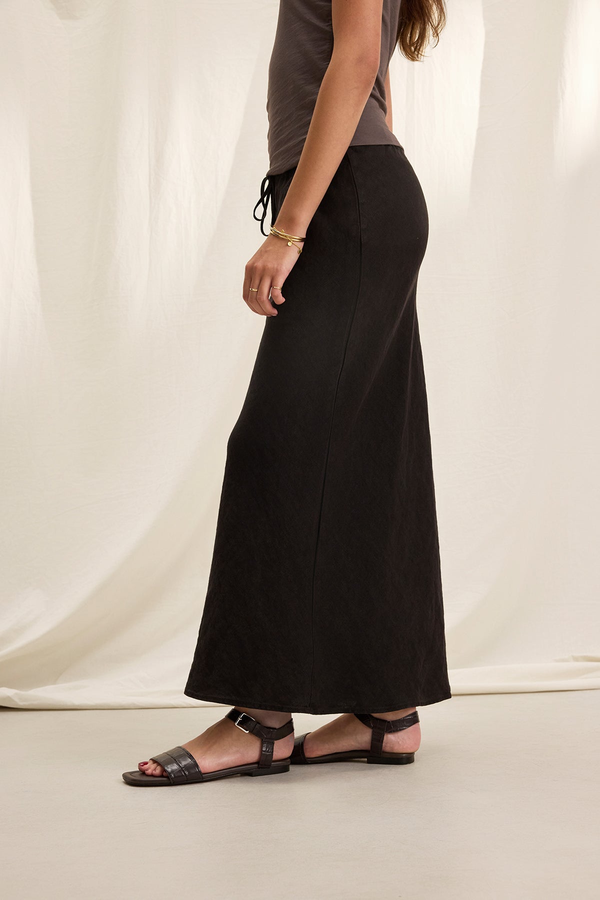 Side view of a person wearing a gray top, the BEKA HEAVY LINEN SKIRT by Velvet by Graham & Spencer, and black sandals, standing against a light curtain background.-38662914179265