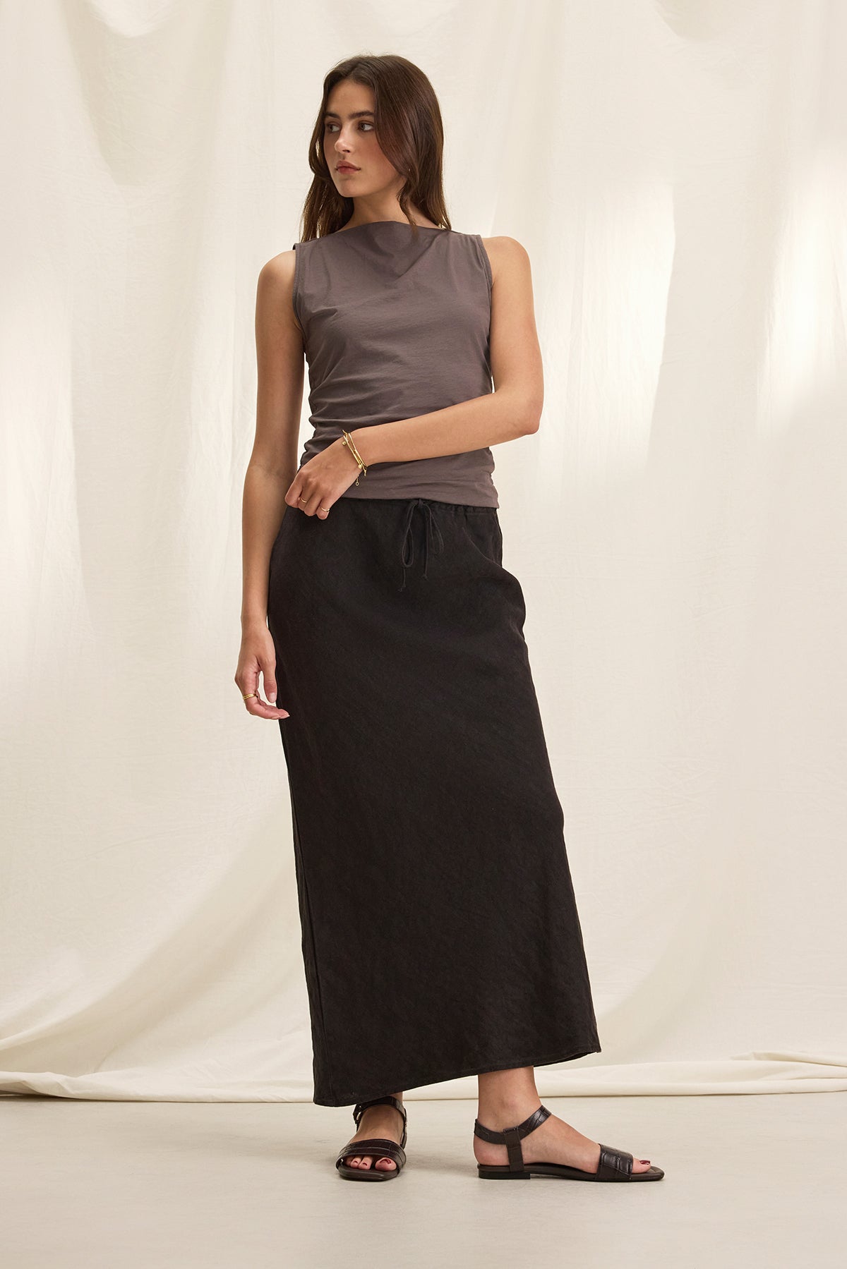 A woman stands against a white backdrop, wearing a sleeveless gray top, the BEKA HEAVY LINEN SKIRT by Velvet by Graham & Spencer with a graceful bias cut, and black sandals.-38662914277569