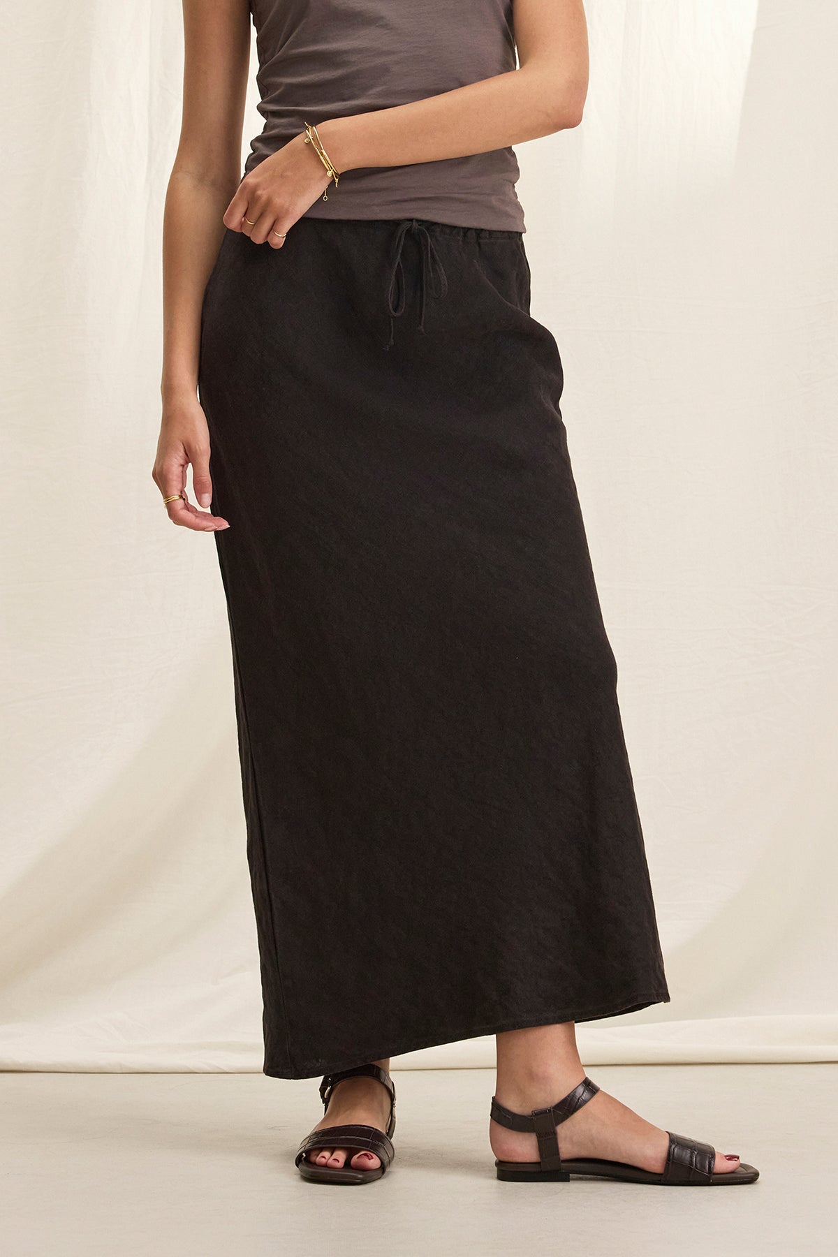 A person stands against a light cream background, wearing a sleeveless gray top, black sandals, and the BEKA HEAVY LINEN SKIRT by Velvet by Graham & Spencer.-38662914244801