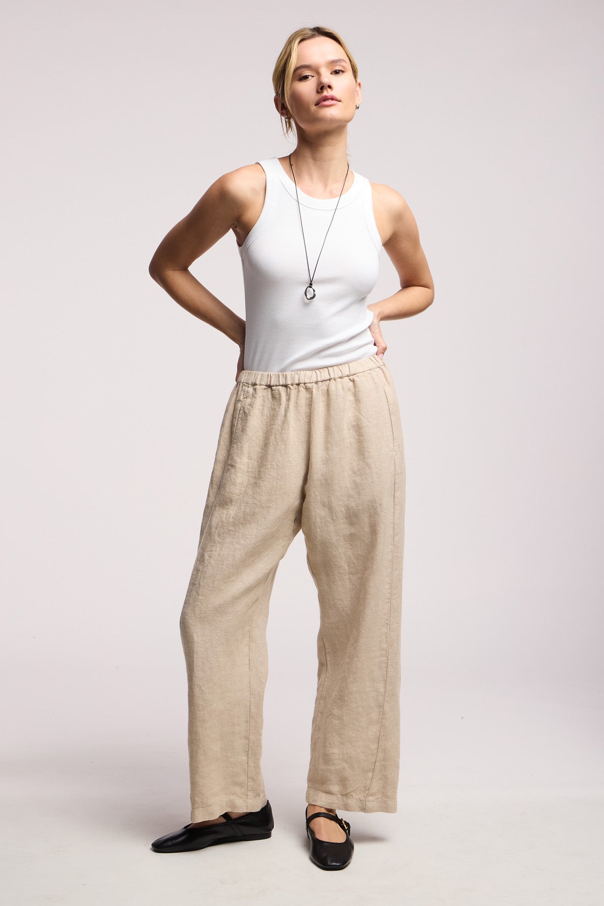 A woman stands against a plain background wearing a white sleeveless top, black flats, a long necklace, and the CELINE HEAVY LINEN PANT by Velvet by Graham & Spencer—wide-leg, cropped pants with an elastic waist.-41371568931009