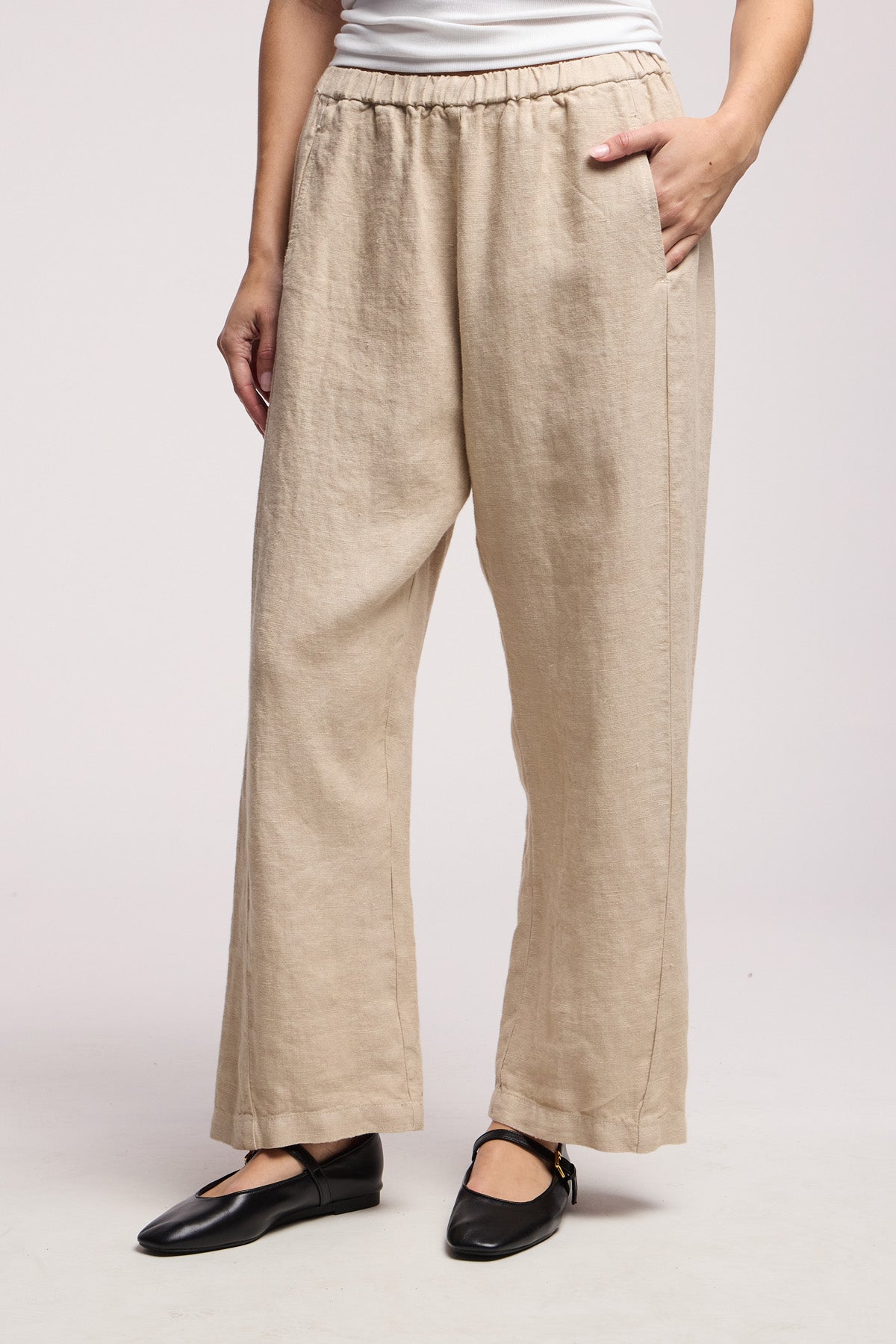 Model wears the CELINE HEAVY LINEN PANT by Velvet by Graham & Spencer—beige, wide-leg, cropped with an elastic waist—styled with a white top and black ankle-strap flats. One hand in her pocket, set against a plain light gray background.-41371568832705