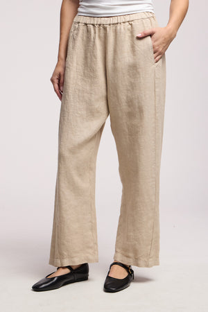 Model wears the CELINE HEAVY LINEN PANT by Velvet by Graham & Spencer—beige, wide-leg, cropped with an elastic waist—styled with a white top and black ankle-strap flats. One hand in her pocket, set against a plain light gray background.