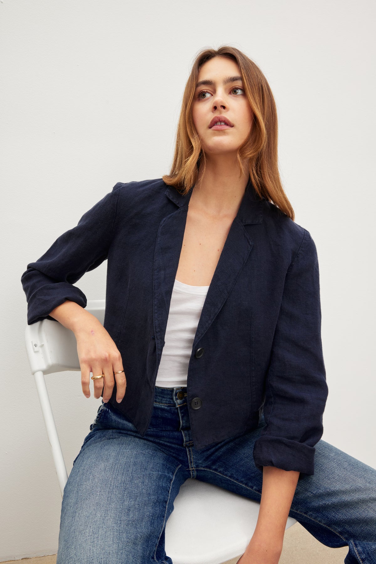   A person with long hair wearing the FINLEY HEAVY LINEN CROPPED BLAZER from Velvet by Graham & Spencer, paired with a white top and blue jeans, showcasing sophisticated styling sits on a white chair against a plain background. 