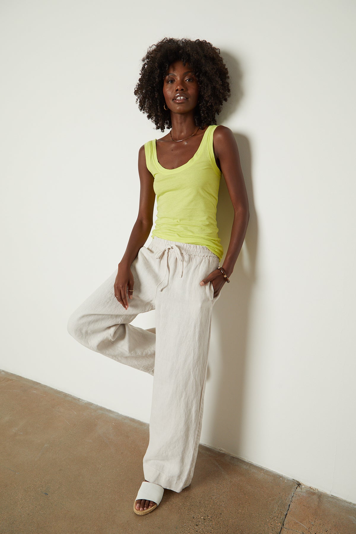   A black woman in a Velvet by Graham & Spencer yellow tank and white pants leaning against a wall. 