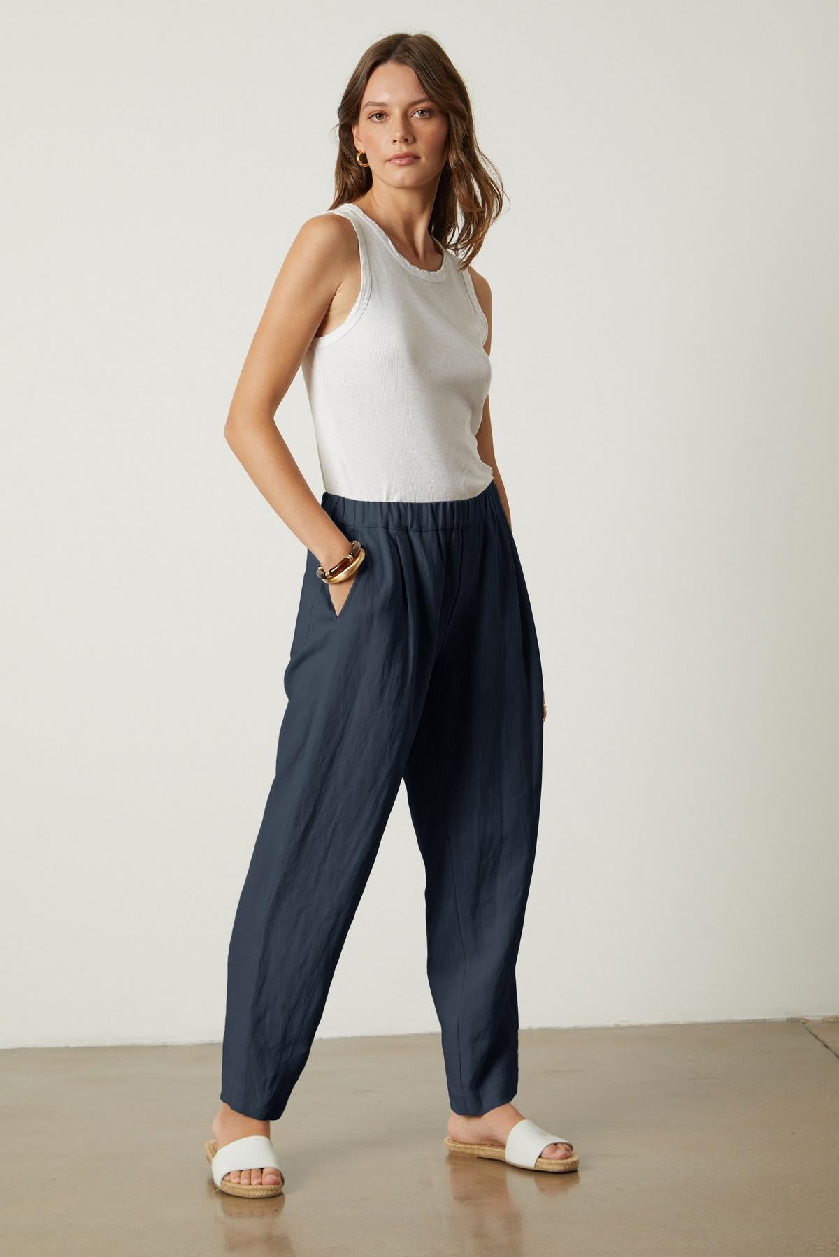  The model is wearing a white tank top and Velvet by Graham & Spencer navy linen trousers, showcasing the drape and texture of the lightweight fabric of the JESSIE HEAVY LINEN PANT. 