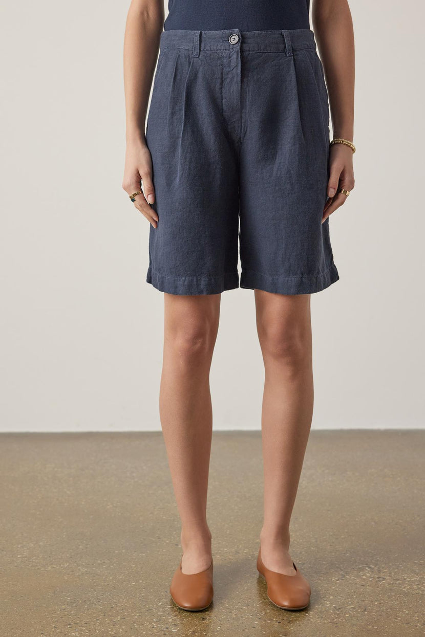 A woman stands wearing Velvet by Jenny Graham's LARCHMONT HEAVY LINEN SHORT and tan loafers, focusing on the lower half of the body against a neutral background.