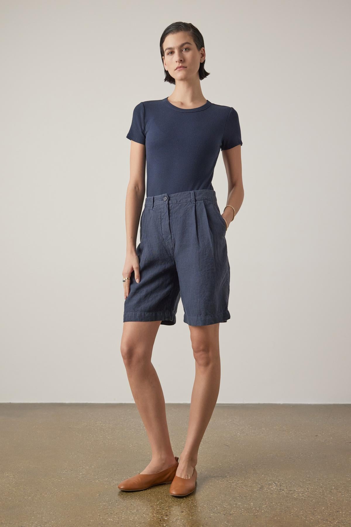 Person standing, wearing a navy blue t-shirt, Velvet by Jenny Graham's dark blue LARCHMONT HEAVY LINEN SHORT, and tan flats, showcasing warm-weather styling against a plain background.-37629303259329