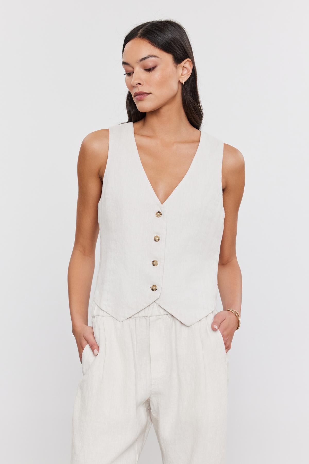   A woman in a beige sleeveless v-neck top with a Velvet by Graham & Spencer MYLIE HEAVY LINEN VEST and matching pants stands against a plain background, looking to the side. 