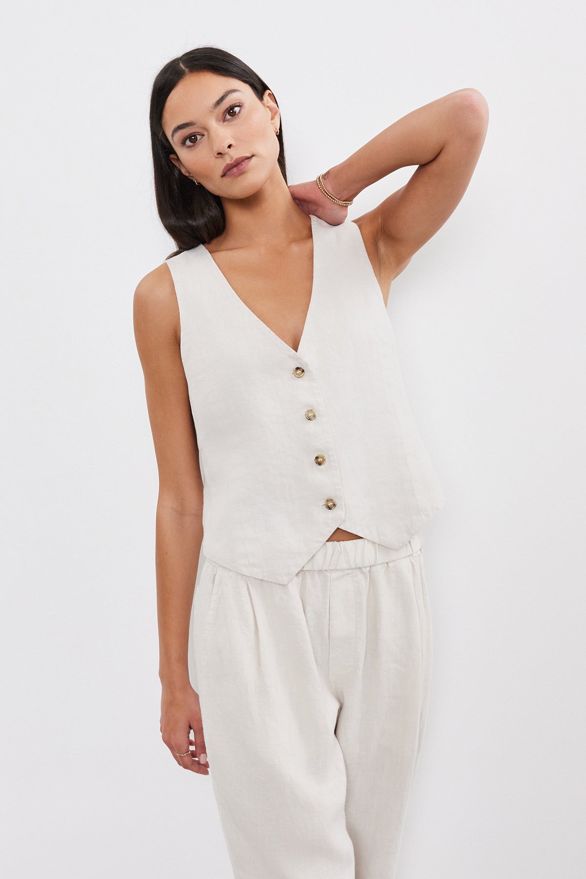   A woman in a light beige sleeveless top and matching pants, topped with a Velvet by Graham & Spencer MYLIE HEAVY LINEN VEST featuring a four-button front, poses against a white background, looking at the camera. 