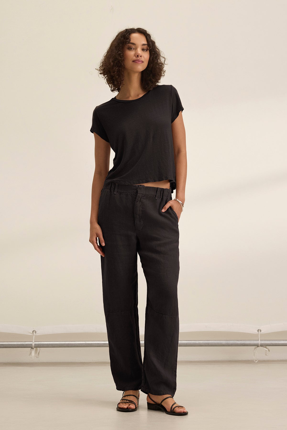 A woman with short curly hair stands indoors wearing Velvet by Graham & Spencer’s TAYLOR HEAVY LINEN PANT in black, styled with a black tailored t-shirt and sandals. The pants are loose-fitting with side seam pockets; she has one hand in her pocket.-39144939749569