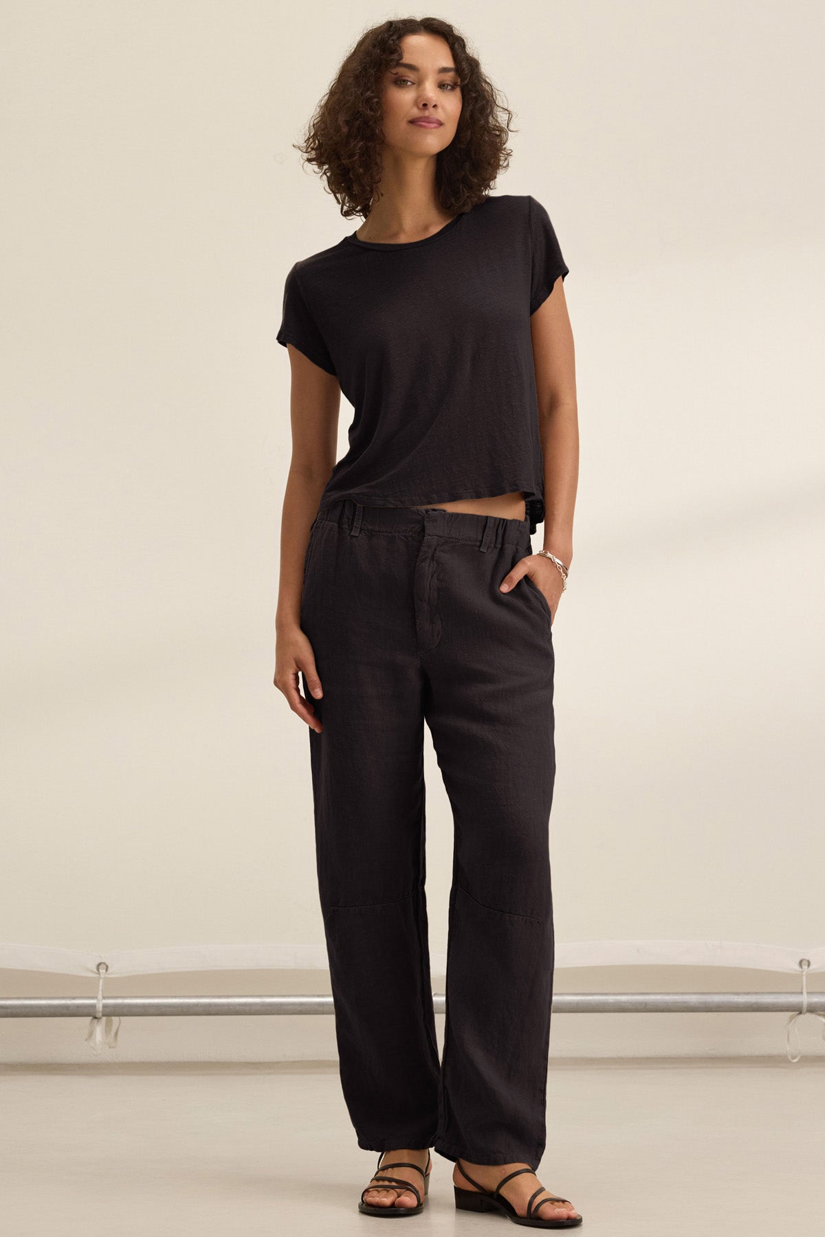 A person stands indoors against a neutral background wearing loose black TAYLOR HEAVY LINEN PANT by Velvet by Graham & Spencer, a tailored fit short-sleeve black t-shirt, and black sandals.-41235234980033