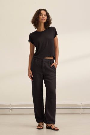 A woman with short curly hair stands indoors wearing Velvet by Graham & Spencer’s TAYLOR HEAVY LINEN PANT in black, styled with a black tailored t-shirt and sandals. The pants are loose-fitting with side seam pockets; she has one hand in her pocket.