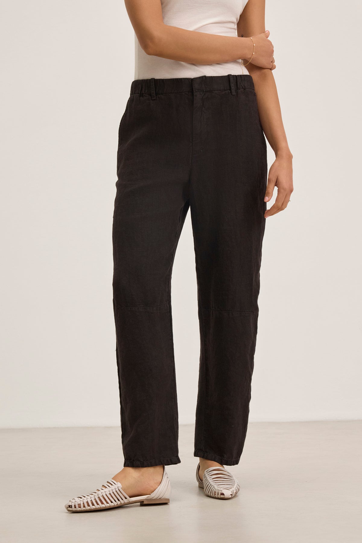A person wears Velvet by Graham & Spencer's TAYLOR HEAVY LINEN PANT in black with a white sleeveless top and white woven flats, standing against a plain background.-39111849738433